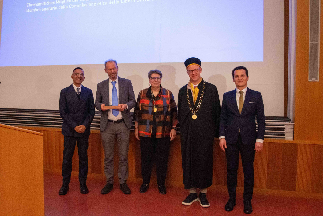 Sechs erwachsene Personen stehen nebeneinander auf einer Bühne in einem Hörsaal. Sie tragen formelle Kleidung; eine Person trägt eine akademische Robe mit Amtskette, im Hintergrund ist eine Projektionsfläche.