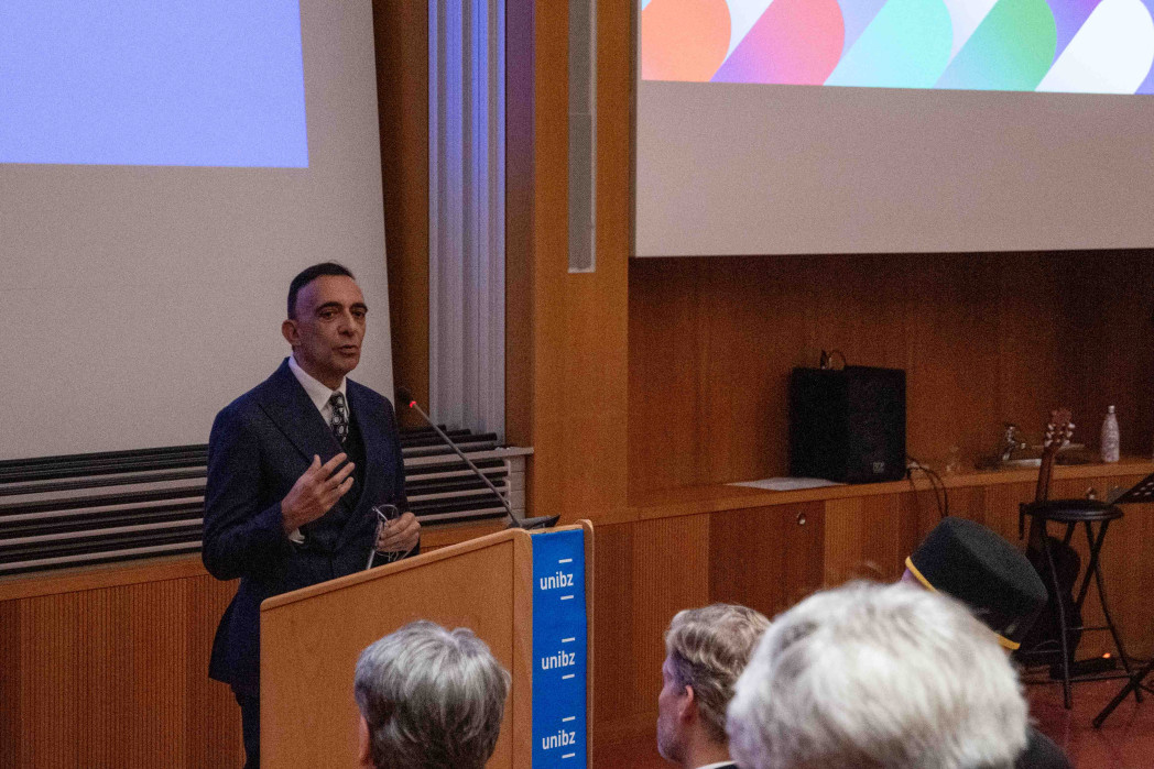 Un uomo in abito parla da un podio in un’aula universitaria, gesticolando con una mano. Davanti a lui è seduto il pubblico; sullo sfondo si vedono pannelli in legno e uno schermo di proiezione.