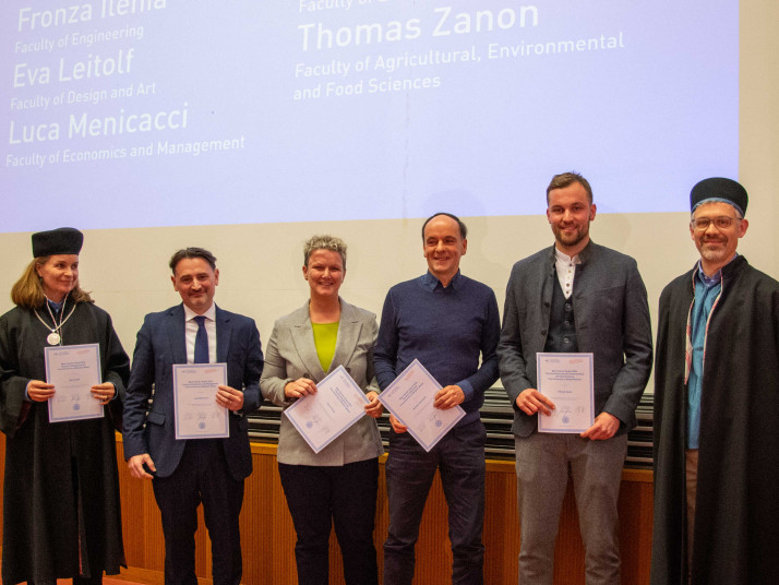 Sette adulti sono in piedi affiancati su un palco di un’aula e tengono dei certificati in mano. Indossano abiti prevalentemente formali; sullo sfondo si vede uno schermo di proiezione con del testo.