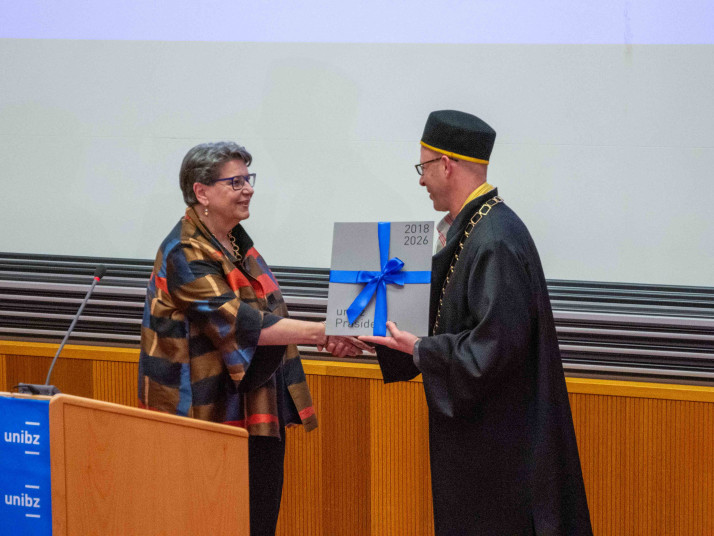 Due persone si scambiano un libro con un nastro blu in un’aula. Una persona indossa una toga accademica con collare cerimoniale; sullo sfondo pannelli in legno e uno schermo di proiezione.