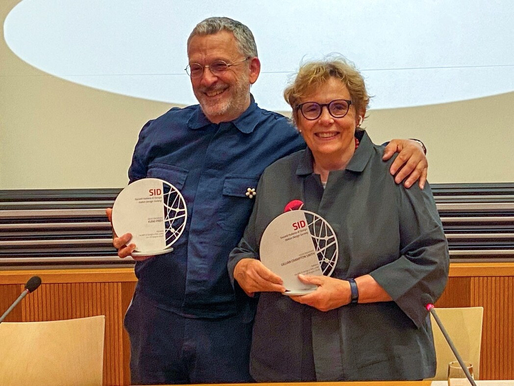 Zwei lächelnde Personen stehen nebeneinander und halten jeweils eine Auszeichnung in den Händen. Eine Person hat den Arm um die Schulter der anderen gelegt. Die Aufnahme entstand in einem Konferenzsaal.