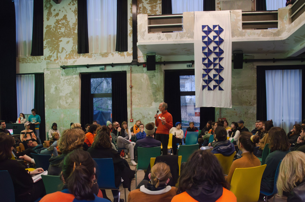 Person spricht vor einer Gruppe sitzender Zuhörer:innen in einem großen Saal mit hohen Fenstern und abgenutzten Wänden; über der Szene hängt ein Banner mit geometrischem Muster.