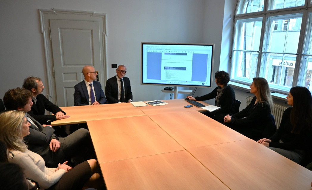 Sette persone in abbigliamento formale sono sedute attorno a un grande tavolo riunioni mentre seguono una presentazione su uno schermo in una sala luminosa.