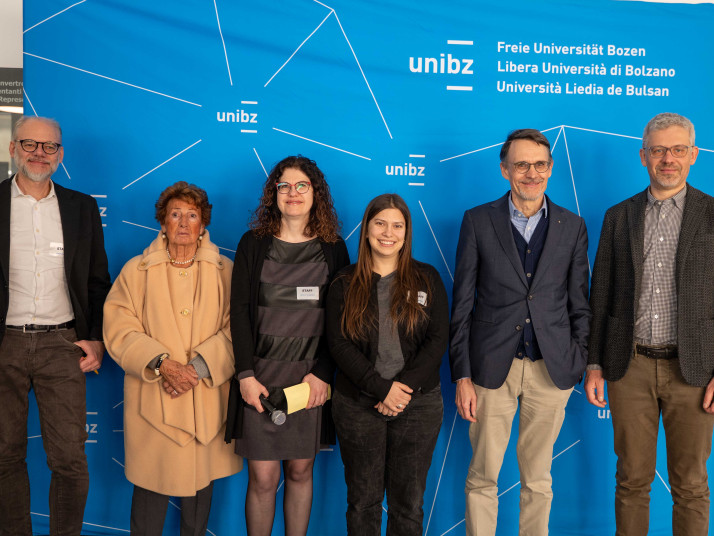 Sei persone stanno in piedi una accanto all’altra davanti a una parete blu con il logo unibz e guardano verso la fotocamera.