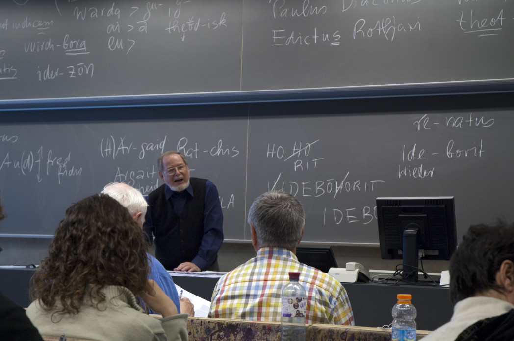Un professore parla agli studenti in un’aula universitaria, davanti a una lavagna con appunti scritti a mano.