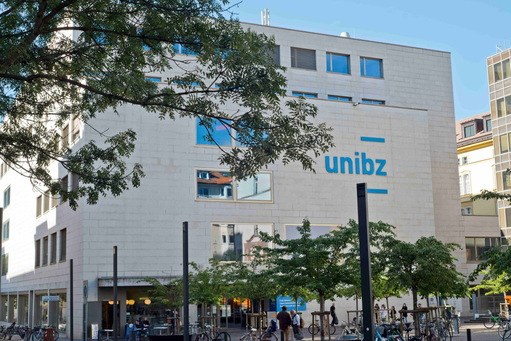 Vista esterna del campus principale della Libera Università di Bolzano a Bolzano: edificio universitario moderno con il logo unibz, alberi e biciclette nella piazza antistante.
