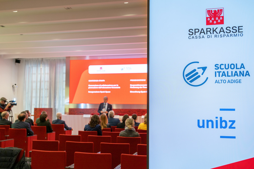 Sala conferenze con pubblico; davanti un uomo parla davanti a una presentazione. A destra un display con i loghi di Sparkasse, Scuola Italiana Alto Adige e unibz.