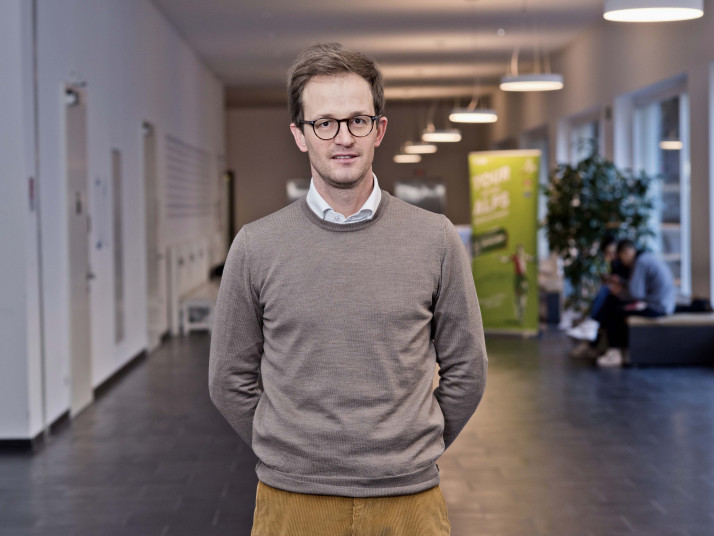 Junger Mann in lässiger Kleidung steht lächelnd in einem Innenraum mit unscharfem Hintergrund.