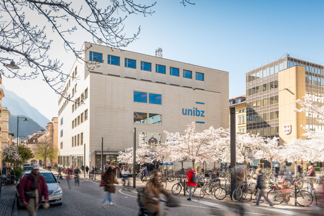 Straßenszene vor modernen Gebäuden mit vielen Fahrrädern und blühenden Bäumen, Menschen gehen und fahren vorbei.