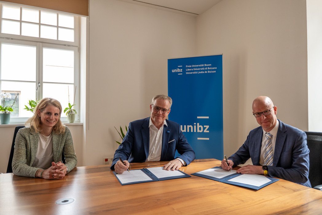 Drei Personen sitzen an einem Tisch; zwei Männer unterzeichnen Dokumente, während eine Frau daneben lächelt. Im Hintergrund steht ein Banner der Freien Universität Bozen.