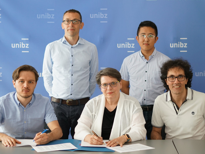 Fünf Personen sitzen und stehen vor einem blauen Hintergrund mit „unibz“-Logo; drei sitzen an einem Tisch mit Dokumenten und Stiften, zwei stehen dahinter.