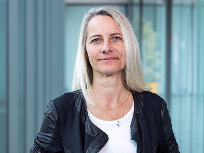 Porträt einer Frau mit schulterlangem Haar und neutralem Gesichtsausdruck vor einem unscharfen Hintergrund aus Glasfenstern; sie trägt eine Jacke über einem Oberteil und eine feine Halskette.