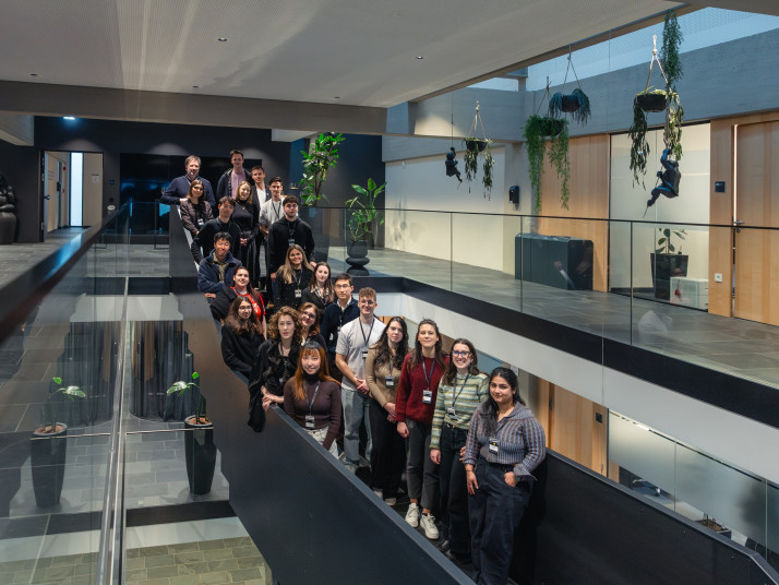 Gruppenfoto von etwa 20 Personen mit Namensschildern, die auf einer Galerie in einem modernen Gebäude mit Glasgeländer, Holztüren und hängenden Pflanzen stehen und in die Kamera blicken.