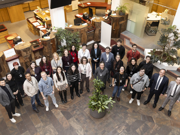 Gruppenfoto von etwa 25 Personen mit Namensschildern, die in einem halbkreisförmigen Foyer mit Holztheken und Pflanzen stehen und in die Kamera lächeln.
