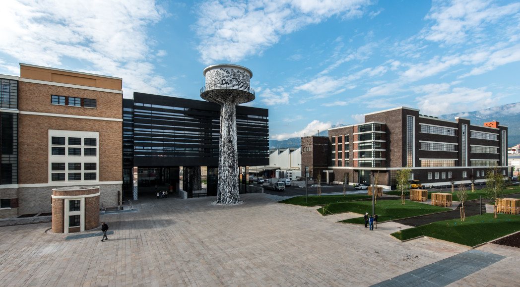 Moderner Universitätscampus mit mehreren Gebäuden aus Glas und Ziegel, einem zentralen Platz und einem markanten, turmartigen Brunnen.