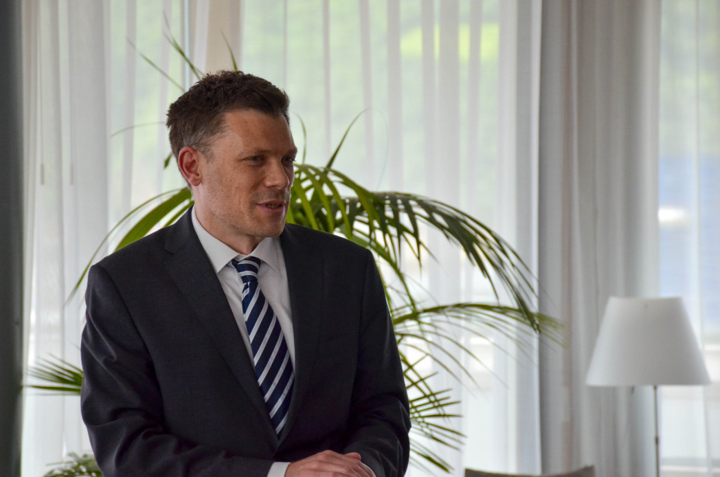 Mann in dunklem Anzug und weißem Hemd blickt in die Kamera, moderne Büroumgebung im Hintergrund