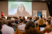 Vielen Menschen sitzen in einem Saal und hören einer Referentin zu. Auf der Bühne ist ihr Abbild projiziert.