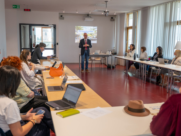 A man gives a presentation to a group of people seated around a U-shaped table in a bright conference room.
