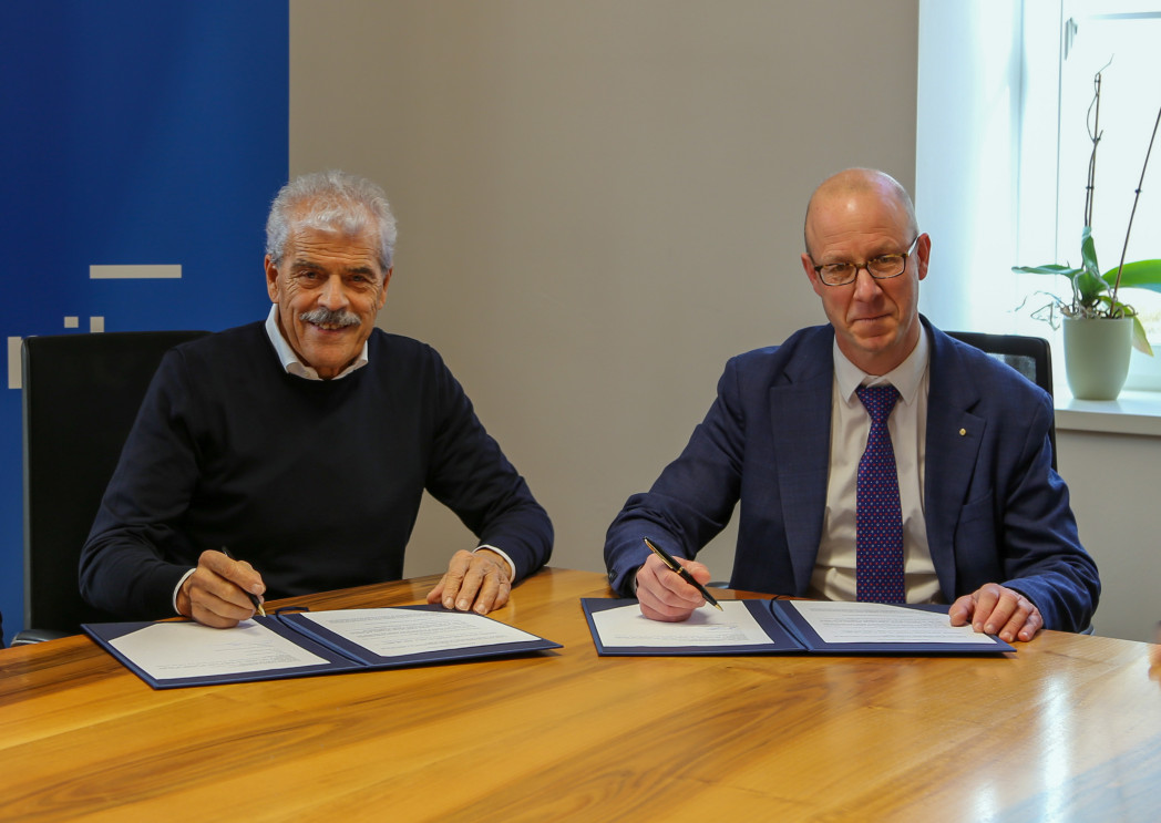 Zwei Männer in Geschäftskleidung unterzeichnen Dokumente an einem Holzkonferenztisch.