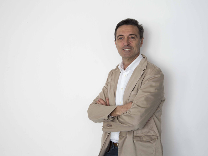 Uomo con le braccia incrociate davanti a una parete bianca guarda verso la fotocamera.