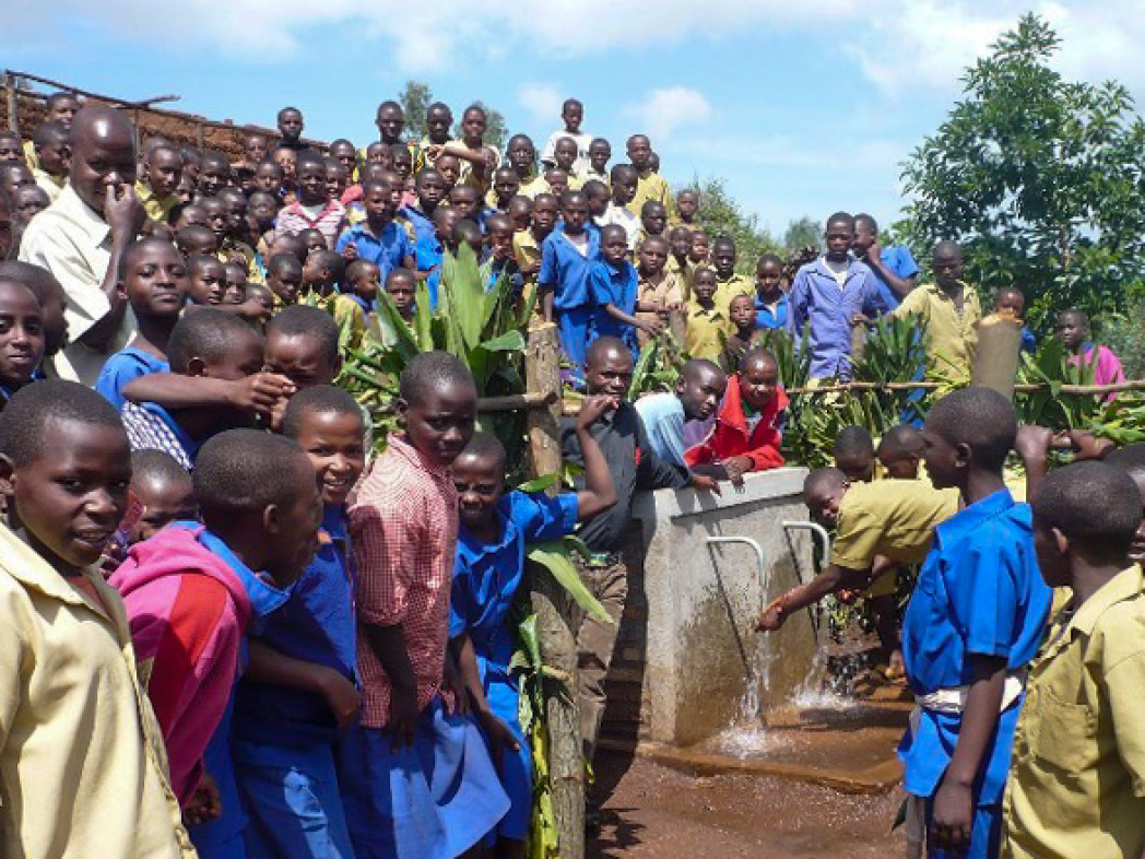 Gruppo di bambini riuniti all’aperto attorno a una pompa d’acqua da cui scorre acqua in un contenitore.
