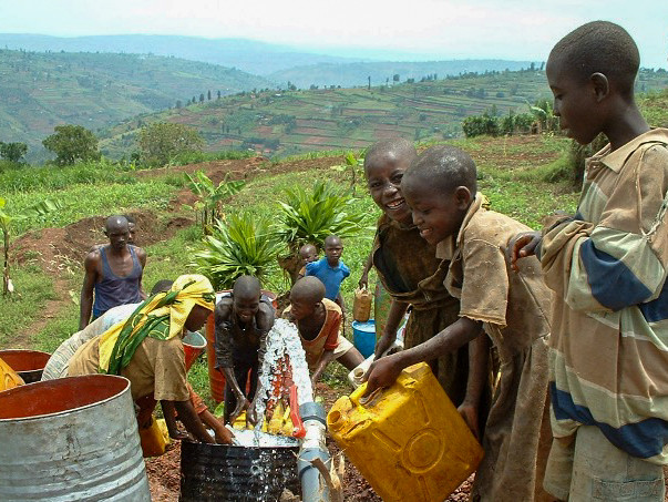 Bambini riempiono taniche d’acqua da una tubazione in un paesaggio rurale collinare.