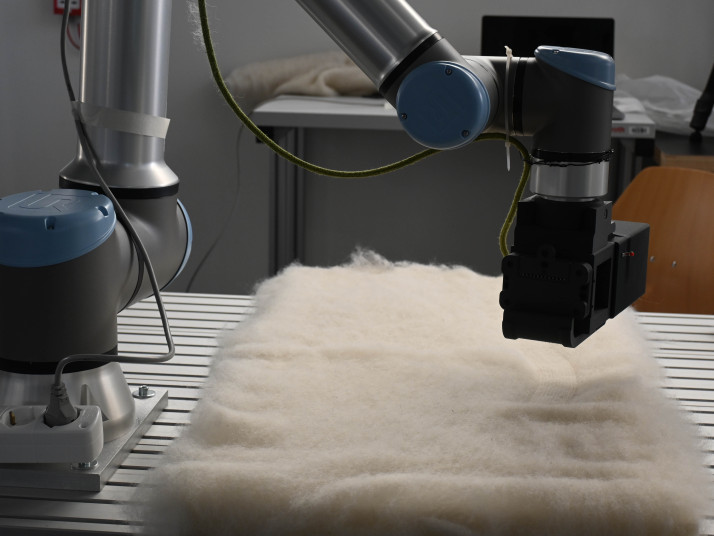 Zwei kollaborative Roboterarme arbeiten über einer hellen Filzmatte auf einem Arbeitstisch in einer Laborumgebung.