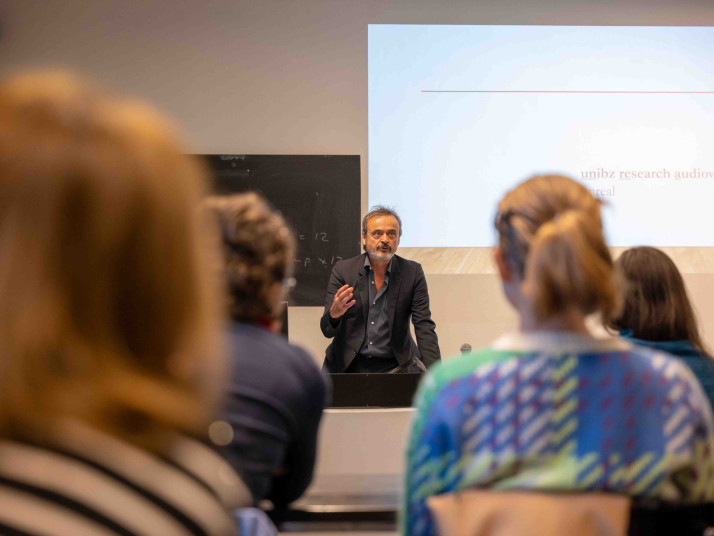 Eine Person spricht vor Publikum in einem Hörsaal der Freien Universität Bozen. Im Hintergrund wird eine Folie zum audiovisuellen Forschungslabor der Universität projiziert