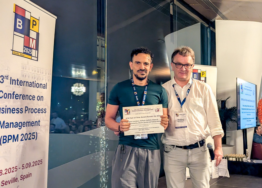 Due uomini in piedi davanti a banner della conferenza BPM, uno in maglietta blu e uno con occhiali in camicia bianca, tengono insieme un certificato di riconoscimento.