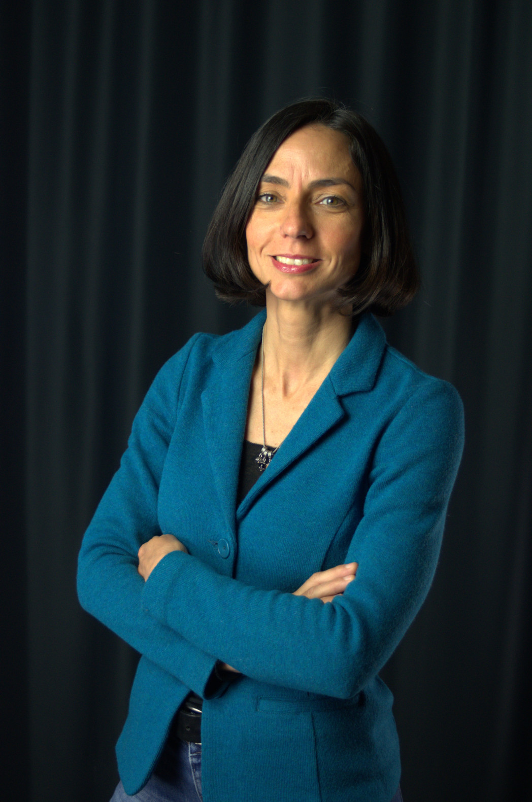 Donna con capelli castani corti, in giacca turchese, in piedi a braccia conserte davanti a uno sfondo scuro.