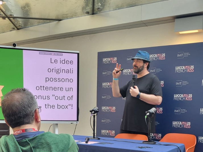 Uomo con cappello tiene un intervento davanti a pubblico e schermo con testo italiano.