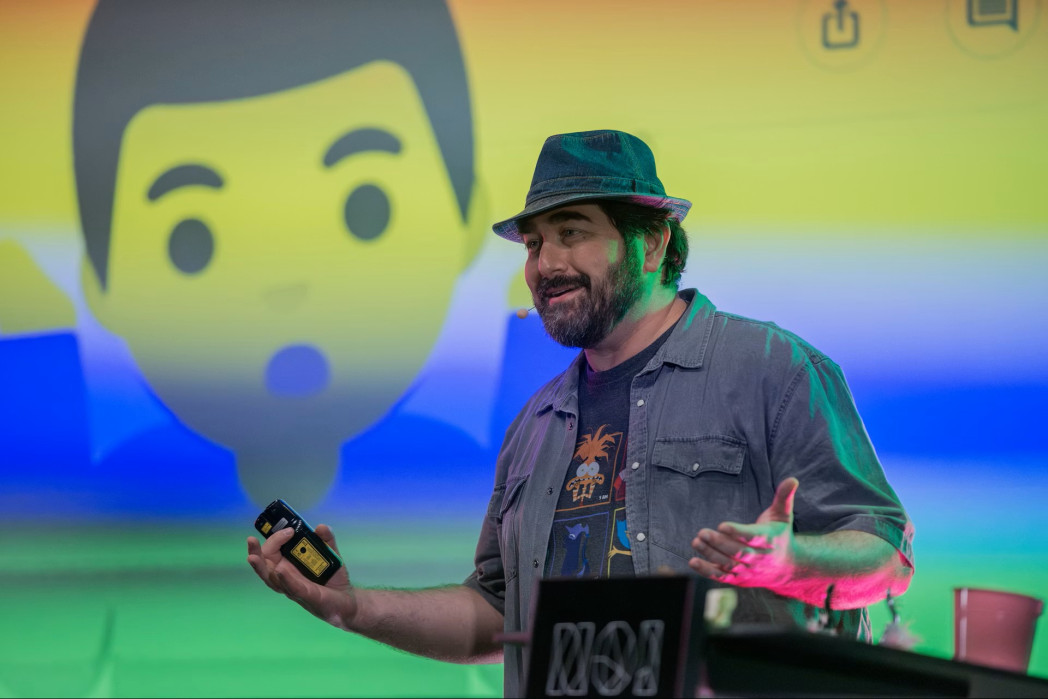 Uomo con barba e cappello parla sul palco davanti a una grafica colorata con un’emoji.