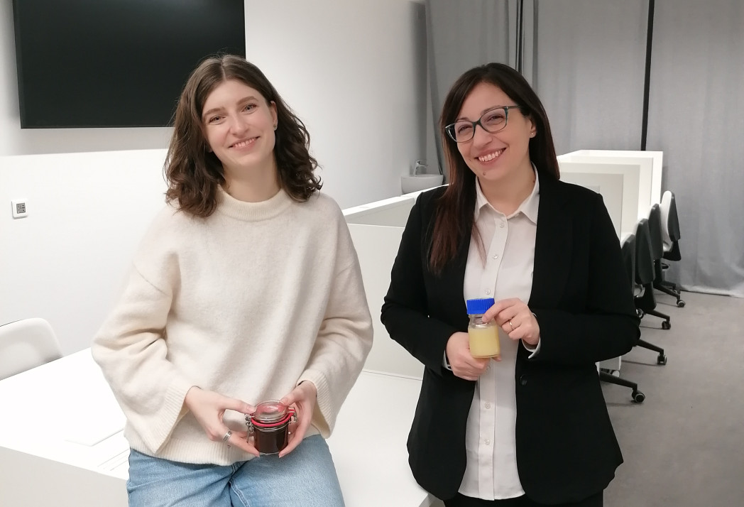 Due donne sorridenti sono in piedi in un ufficio luminoso e tengono ciascuna un piccolo barattolo.