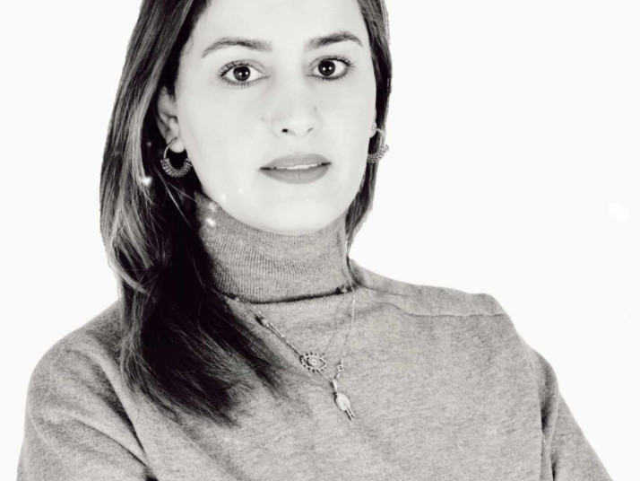 Portrait of a woman with shoulder-length hair looking directly at the camera, wearing a turtleneck sweater and jewelry against a light background.