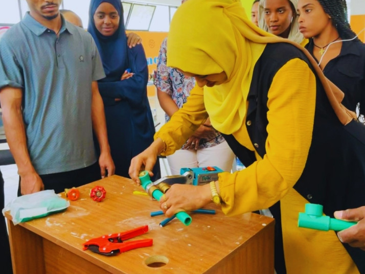 A group of young people stand around a table watching a woman in a yellow headscarf use a tool on a plastic pipe in a workshop setting.