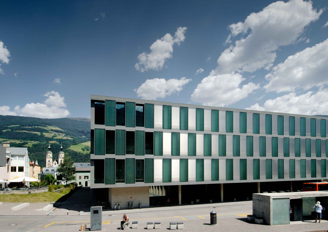 Modernes Universitätsgebäude mit grün-weißer Fassade vor bewaldeten Bergen und blauem Himmel.