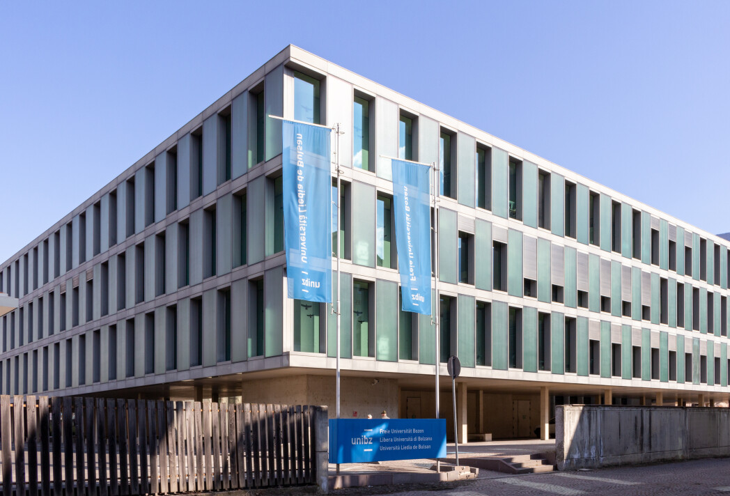 Modernes, vierstöckiges Universitätsgebäude mit großer Glasfassade und zwei unibz-Fahnen vor dem Eingang unter klarem Himmel.